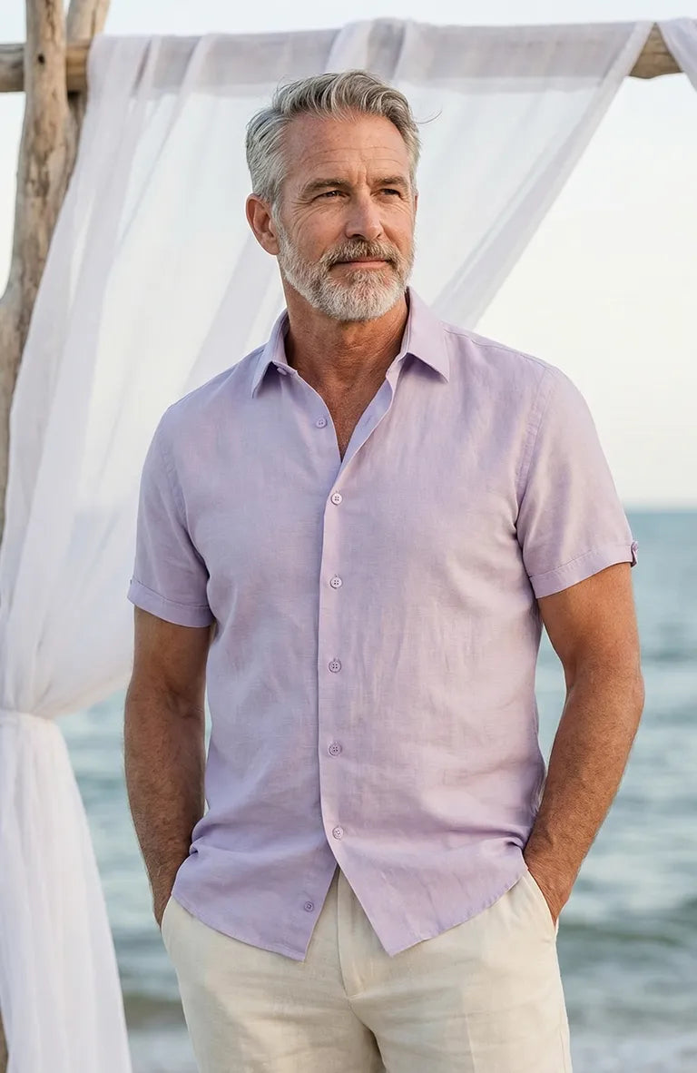Man wearing a Beckford Men’s Linen Shirt in Violet and beige pants by a white curtain with a blurred beach background