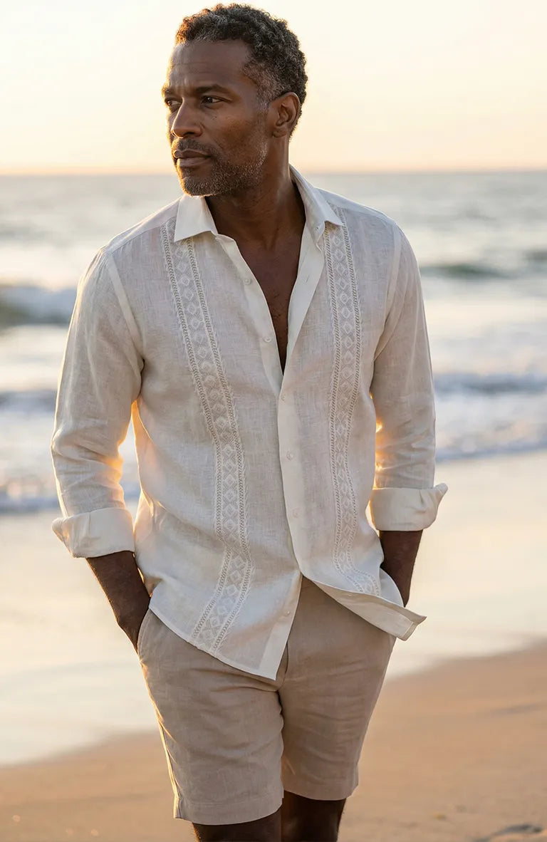 Man wearing a light-colored Guayabera Shirt and shorts on a beach at sunset.