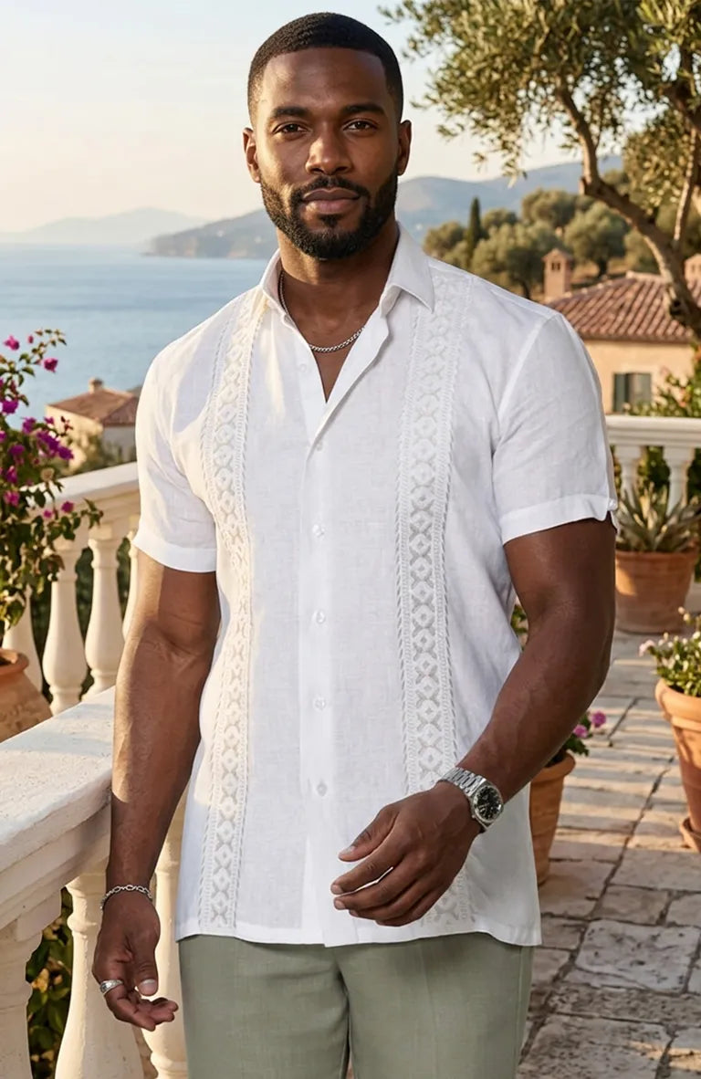 Man wearing a white embroidered Guayabera Shirt and light green pants standing on a balcony with a scenic view.
