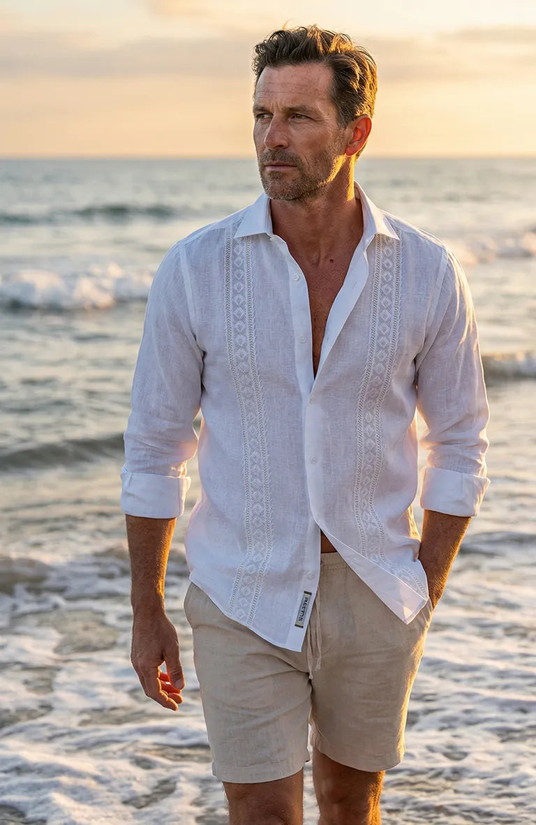 Man wearing a white Guayabera Shirt  and beige shorts standing on a beach at sunset.
